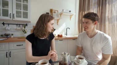 Young couple enjoys drinking hot tea sitting at glass kitchen table and talking. Woman turns to camera to continue online communication from third party to POV