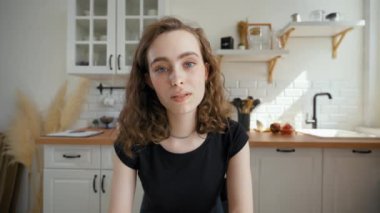 Young woman talks looking in camera and drinking tea against countertop in kitchen. Lady turns to man sitting at glass table from POV to third party