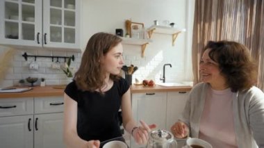 Daughter and mother enjoy drinking tea sitting at glass table in kitchen. Young woman turns to camera to continue talking on video call from third party to POV