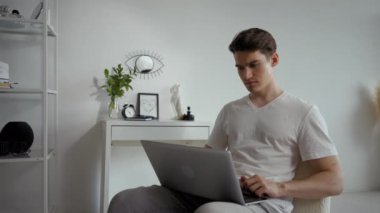 Young man sits on chair against white wall working on laptop from stylish room. Male freelancer types report of new project using modern electronic device