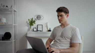 Focused employee looks at screen of laptop typing text. Young man works online using modern electronic device put on laps sitting on chair in living room