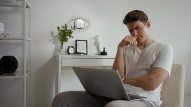 Man feels tired listening to boring online lecture via laptop. Young sleepy student sits on chair in living room yawning and covering mouth with hand
