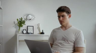 Young employee talks with colleague on video call via notebook put on laps. Man discusses new project sitting on chair against white wall in living room closeup