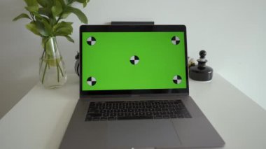 Man sits at white desk talking on video call via laptop near glass vase. Modern electronic device with green chromakey screen on office place to insert