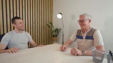 Young businessman talks to elderly client discussing new joint project. Man sits at desk in office turning to camera to summarize from third party to POV