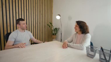 Young man talks to elderly woman applicant sitting at desk in modern office. Employer conducts job interview turning to camera from third party to POV