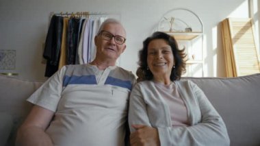 POV of senior grey-haired man and woman discussing funny moments on video call via webcam with friends. Happy couple talk sitting on couch at home closeup