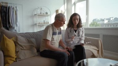 Lady doctor in white coat prescribes medications for elderly patient at home. Specialist shows treatment plan to senior man sitting on sofa in living room