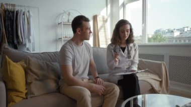 General practitioner explains treatment plan to young patient sitting on couch in living room at home. Doctor in white coat takes care of health of man