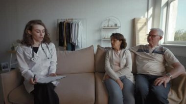Elderly couple talks to general practitioner sitting on couch at home. Senior man and woman turn to camera telling about health condition from third party to POV