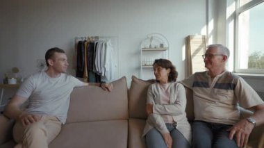Adult son talks to elderly parents sitting on sofa in room. Senior man and woman turn to camera to continue conversation on video call from third party to POV