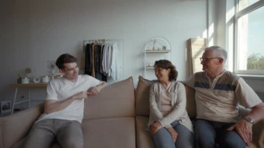 Adult son discusses funny story with elderly parents sitting on sofa in living room. Senior man and woman laugh getting back to video call from third party to POV