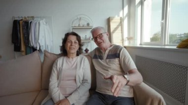 Elderly couple talks on camera discussing fun situation with friends. Senior man and woman turn to talk to son student sitting on sofa from POV to third party