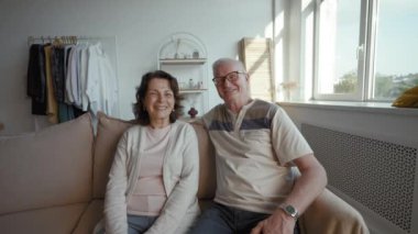 Happy senior man and woman enjoy conversation on video call with friends. Elderly couple turns to talk to daughter sitting on sofa from POV to third party