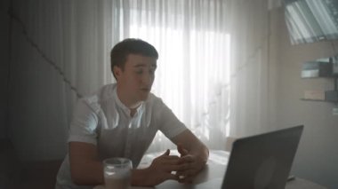 Young man talks on video call with girlfriend via laptop sitting at table against bright window. Guy enjoys online communication at home closeup
