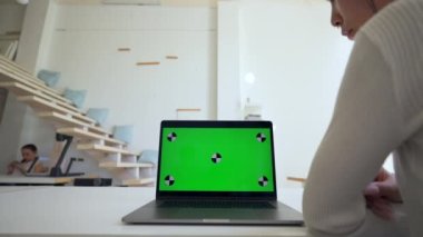 Woman watches online conference sitting at desk in office. Modern laptop with green chromakey screen at workplace ready for tracking closeup