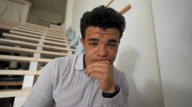 POV of young student feeling tired listening to online teaching courses on webcam. Bored man sits on wooden stairs in living room closeup