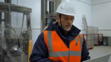 Engineer with grey beard uses laptop looking around premise of warehouse. Mature man in white hardhat stands in storage terminal closeup slow motion