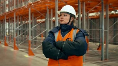 Female engineer with crossed arms stands against empty racks in warehouse. Serious woman in professional uniform turns to camera closeup
