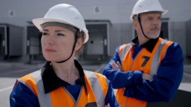 POV of woman and man in uniform standing with crossed arms against warehouse with loading docks. Engineers wear white hardhats looking in camera closeup