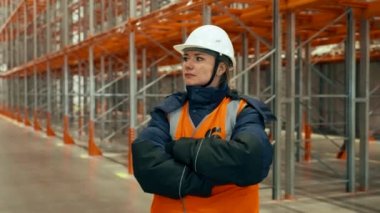 Woman in white hardhat stands with crossed arms against empty metal racks in warehouse. Female engineer turns to camera smiling closeup
