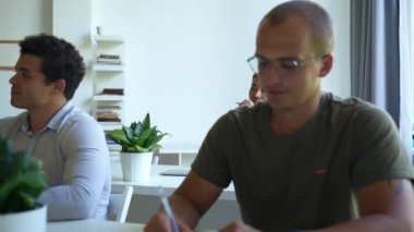 Focused colleagues listen to mentor at teaching conference sitting at desks with pens and paper copybooks. Young man in glasses writes notes closeup