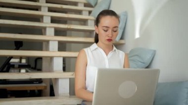 Young woman works on laptop turning to camera and smiling. Female freelancer sits on wooden stairs at home typing article on modern device