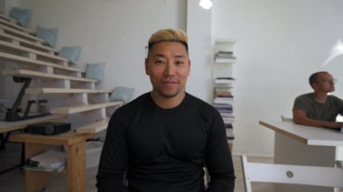 Asian blond man enjoys talking via video call with girlfriend at work break in office. Young guy turns to greet bearded colleague coming to sit from POV to third party
