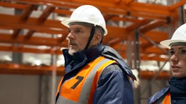 Experienced engineers discuss working issues walking near empty metal racks in warehouse. Man and woman in white hardhats talk closeup slow motion