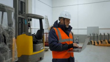 Engineer in professional uniform examines premise of warehouse holding laptop. Bearded man uses device standing in terminal closeup slow motion