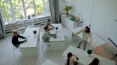 Young employees listen to mentor sitting at white desks in big audience. Man with long hair conducts business training upper angle view slow motion