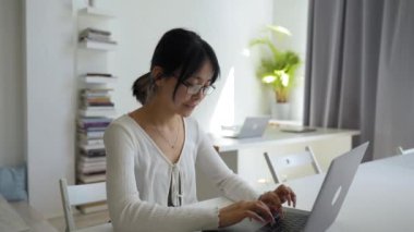 Asian woman in glasses types report of new project on modern laptop. Young employee sits at desk in office turning to camera smiling closeup