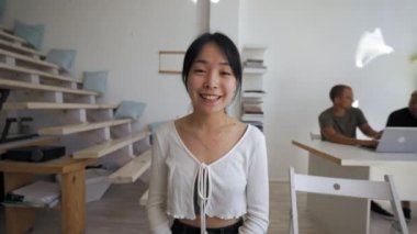 Asian woman waves hello to camera talking to best friend at work break in office. Young lady turns to colleague sitting at desk from POV to third party