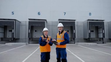POV of man and woman in uniforms standing against open gates of large warehouse complex. Professional engineers with tablet and clipboard look in camera