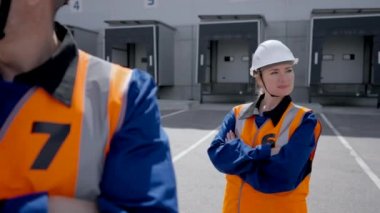 POV of colleagues in uniform standing with crossed arms looking in camera and smiling. Engineers at work against warehouse with open gates closeup