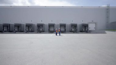 POV of woman and man standing against large industrial building with open gates discussing work. Engineers in uniform turn to approaching camera smiling