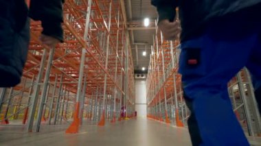 Engineers walk on aisle between empty metal racks in storehouse. Workers in uniform examine premise of storage terminal backside view slow motion