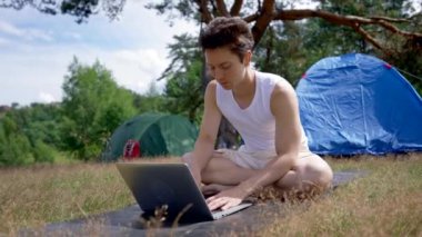 Young man works on laptop typing information while sitting on mat by camp tent. Black-haired freelancer spends time in fresh air working on laptop on sunny day