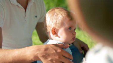Baby boy sits near father pouting cheeks. Father strokes son with large hand. Toddler child examines park territory with careful and amused expression closeup