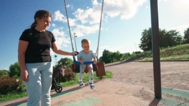 Small toddler boy rides on plastic chain swing smiling and laughing against blue sky. Excited child enjoys walking and playing on playground with mother in park