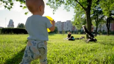 Toddler boy holds ball walking to family at back sunlight. Kid smiles and laughs running on grass on sunny day. Child enjoys going to picnic with parents closeup