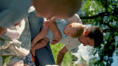 Serious baby boy sits on father laps. Father eats banana holding child spending time on lawn grass. Baby looks wanting to eat delicacies at family park picnic