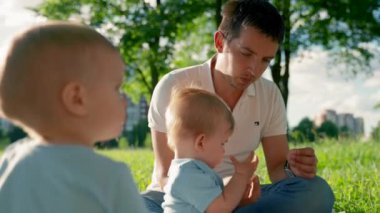 Father gives baby son small piece of banana sitting on picnic grass. Baby boy examines food and takes from father hand. Child looks with serious expression closeup