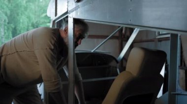 Senior bearded craftsman checks quality of aircraft pilot seat. Aged engineer wearing brown shirt examines airplane for design shortcomings in workshop