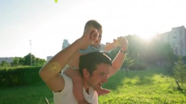 Happy father plays with cute baby girl holding on strong shoulders. Daughter looks around with serious and amused expression on face against park closeup