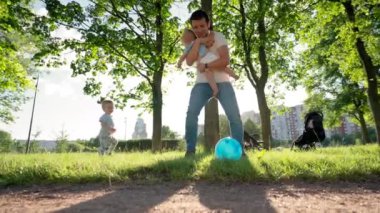 Father holding baby in arms kicks blue ball playing with toddler son. Boy tries to catch ball playing with father. Son enjoys playing ball with father at sunset