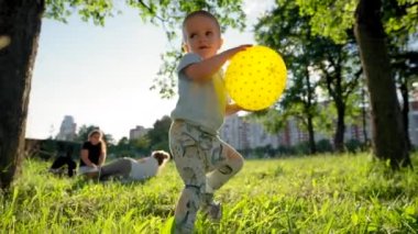 Happy toddler boy plays with yellow ball throwing away. Child walks holding ball in hands against blurry family on picnic. Boy enjoys playing in park closeup