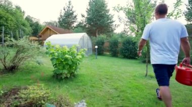 Middle-aged man in t-shirt and shorts goes with watering can to water trees in garden. Mature owner enjoys taking care of groomed garden in morning slow motion