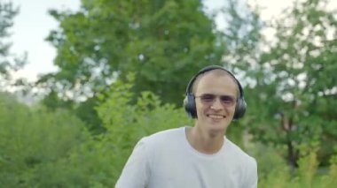 Stylish man waters vegetables dancing in own greenhouse. Middle-aged gardener comes to pavilion of greenhouse listening to loud music in black headphones