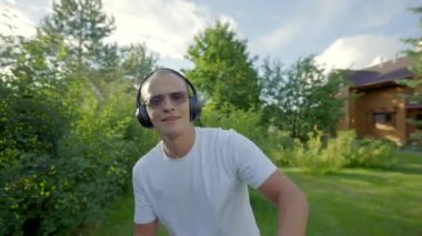 Stylish hipster in headphones and sunglasses enjoys listening to music. Middle-aged gardener comes into greenhouse and waters vegetables dancing closeup